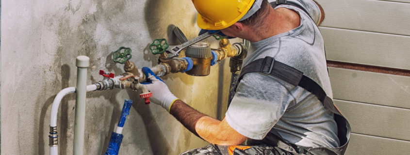Plumber in a yellow hard hat repairs a water pipe with a wrench.